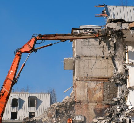 Building demolition with hydraulic excavator. Demolished destructed house ruins in european city on bright sunny day.
