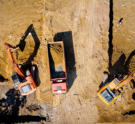 Above top view on two big excavators as they digging, excavating foundation for new residential building and filling few dumper trucks with soil, ground at construction site, project in progress.
